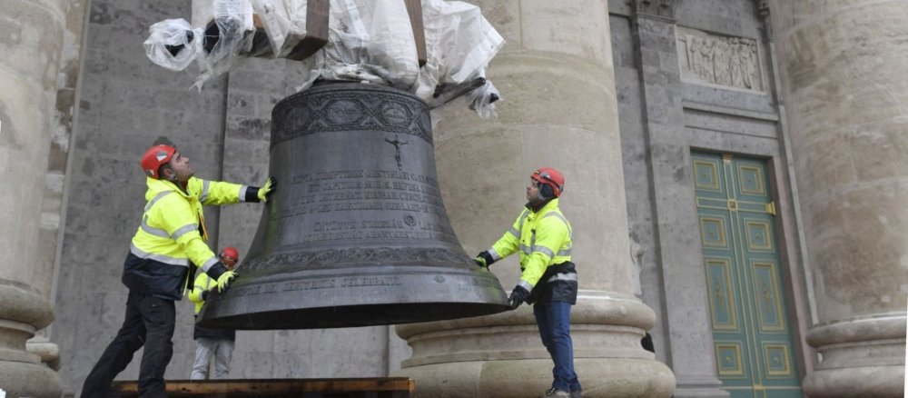 Zwei Männer halten eine Glocke, die an einem Kran hängt.