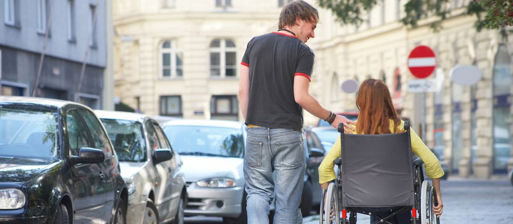 Auf dem Gehweg einer Stadt fährt die Frau im Rollstuhl, ein junger Mann berührt sie an der Schulter.