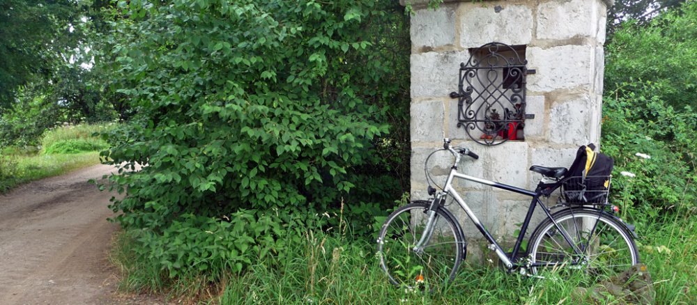 Das Rad lehnt am Bildstock im Gras.