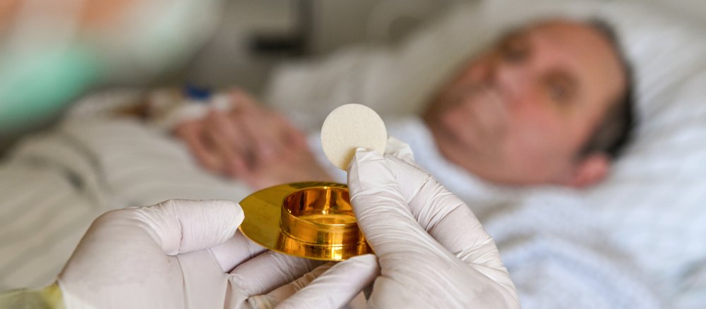 Ein Klinikseelsorger mit Schutzkittel und Atemschutzmaske spendet einem Patienten die Krankenkommunion.