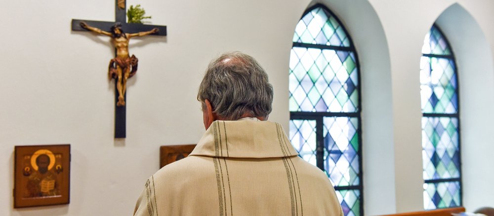Ein Priester steht in einer Sakristei vor einem kreuz und betet.