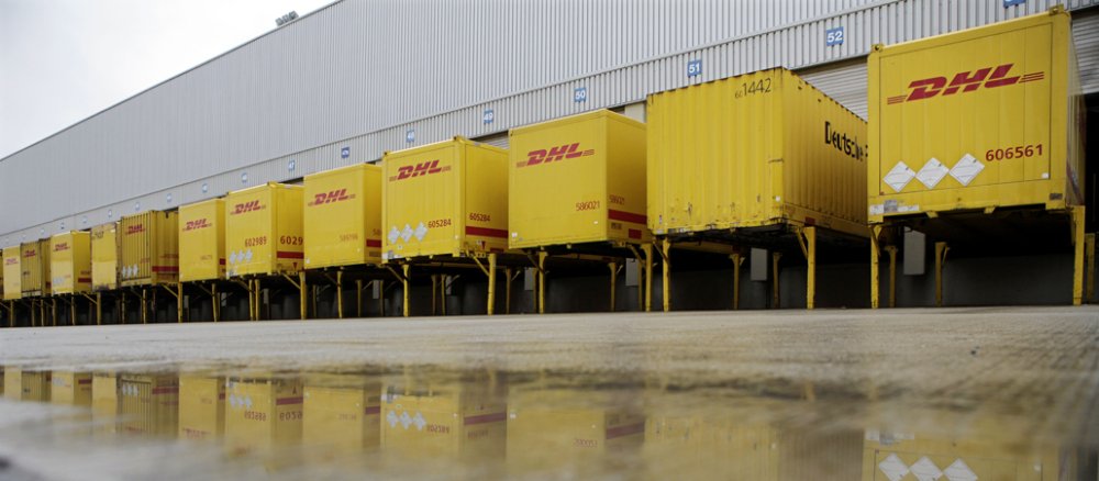 Elf gelbe DHL-Container am Paketzentrum spiegeln sich in einer Pfütze.