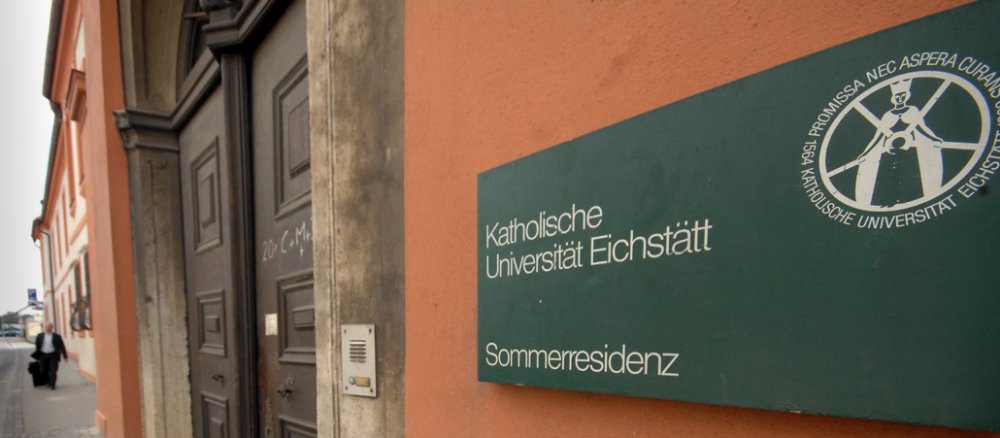 Gebäudefront mit einer dunklen Holztür in Eichstätt. Rechts vor dem Eingang hängt ein großes dunkelgrünes Schild mit der Bezeichnung "Katholische Universität Eichstätt".
