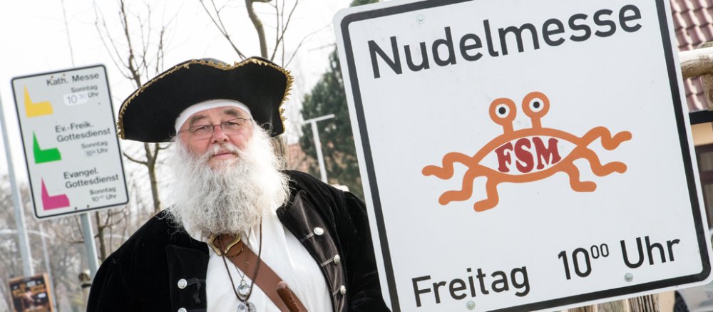 Rüdiger Weida von der "Kirche der fliegenden Spaghettimonster" steht am 05.04.2016 neben seinem Schild "Nudelmesse" in Templin (Brandenburg) und im Hintergrund das Schild der katholischen und evangelischen Kirche zu sehen.