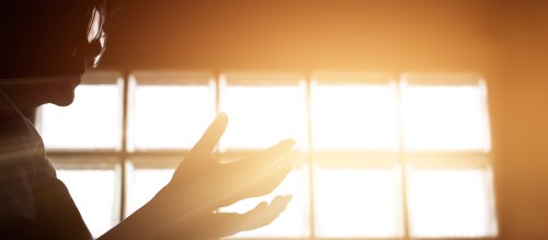 Im Gegenlicht eines Fensters sieht man nur die reflektierenden Brillengläser und die vor der Brust nach oben gehobenen Hände eines Menschen.