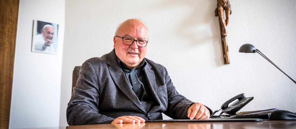 Theo Paul an sitzt an seinem Schreibtisch, im Hintergrund hängt ein Bild von Papst Franziskus