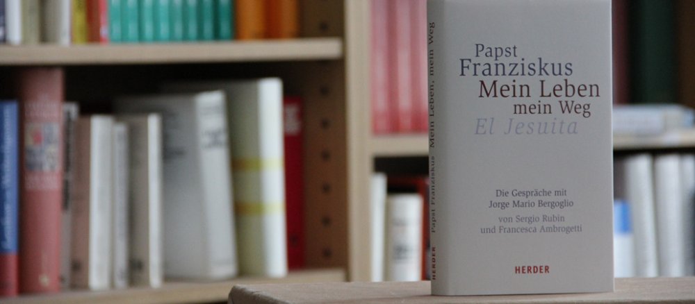 Das in hellem Beige gehaltene Buch steht vor einem Bücherregal.