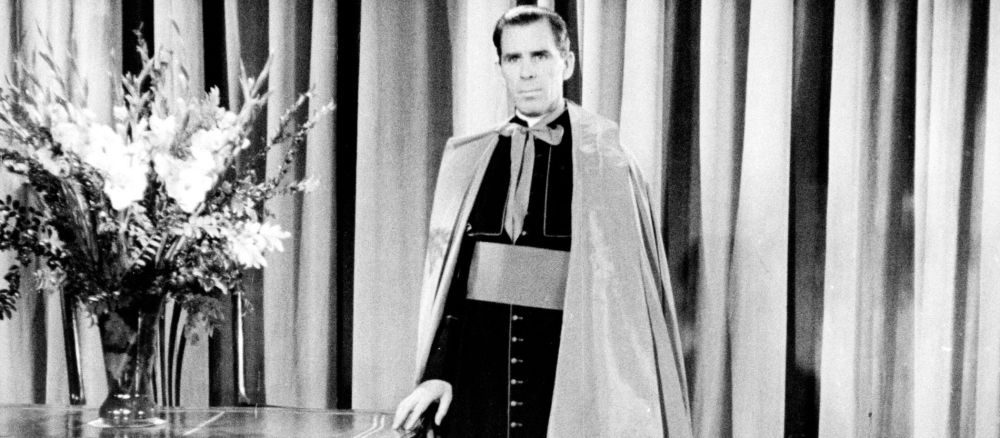 Der Fernsehbischof Fulton Sheen lehnt sich mit einer Hand an einen Tisch an. Er trägt seine Bischofskleidung.