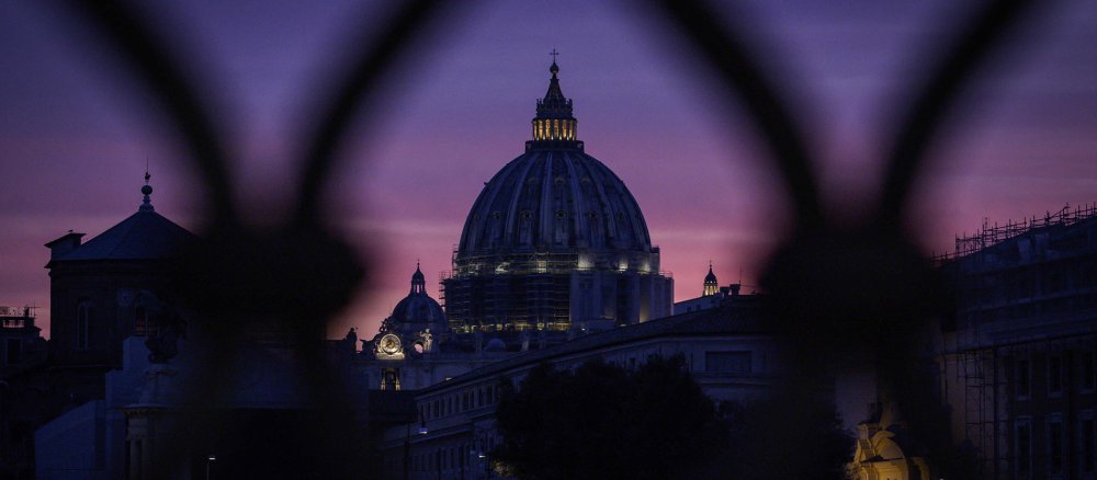 Die Kuppel des Petersdoms, fotografiert durch die Maschen eines Zauns, vor rosa und lila farbenem Abendhimmel