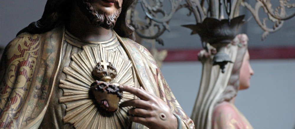 Die Jesus-Statue deutet mit dem Finger auf ihr freiliegendes Herz, das von Dornen umrankt ist und oben brennt und ein Kreuz aufweist.