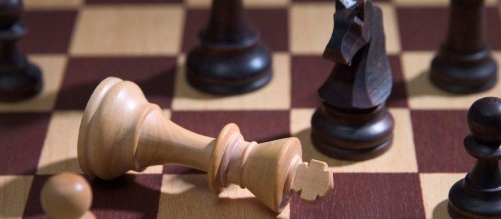 Der weiße König liegt geschlagen auf einem Schachbrett - um ihn herum die Figuren des schwarzen Spielers.