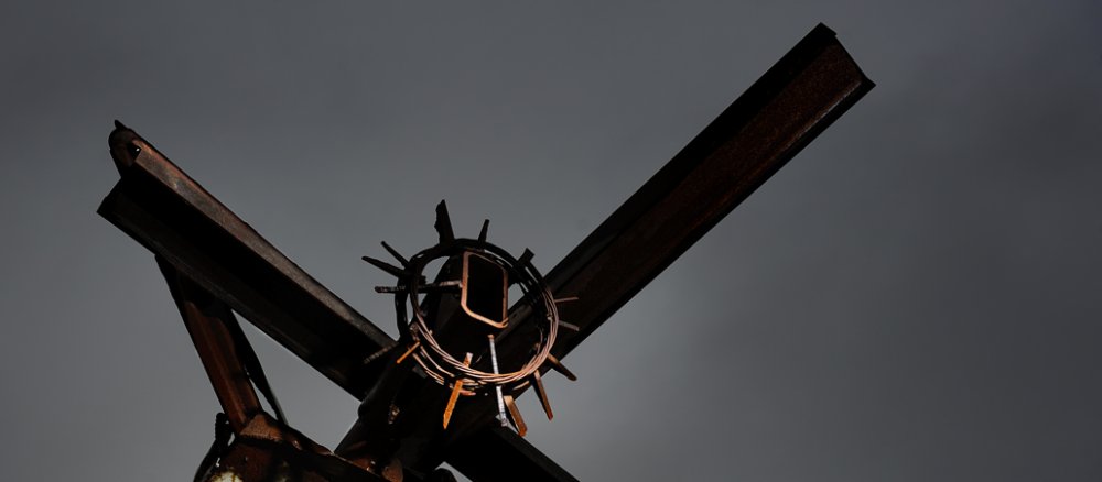 Skulptur aus rostendem Metall zeigt stilisierten Jesus mit Dornenkrone am Kreuz.