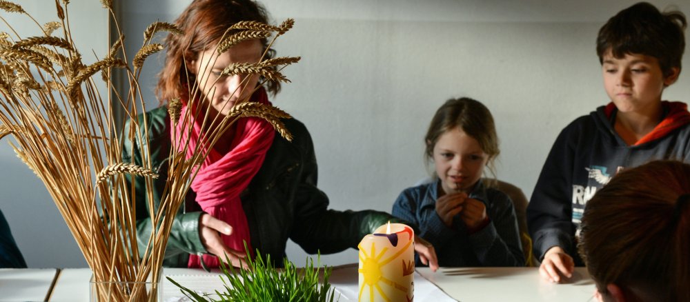 Eine Katechetin spricht mit Kindern. Auf dem Tisch steht ein Glas mit Ähren und eine Kerze.