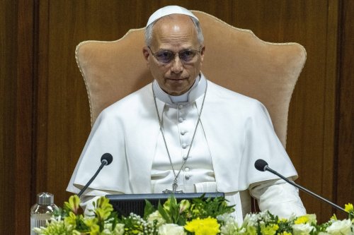 Papst Leo sitzt im weißen Gewand auf einem großen Stuhl vor zwei Mikrofonen