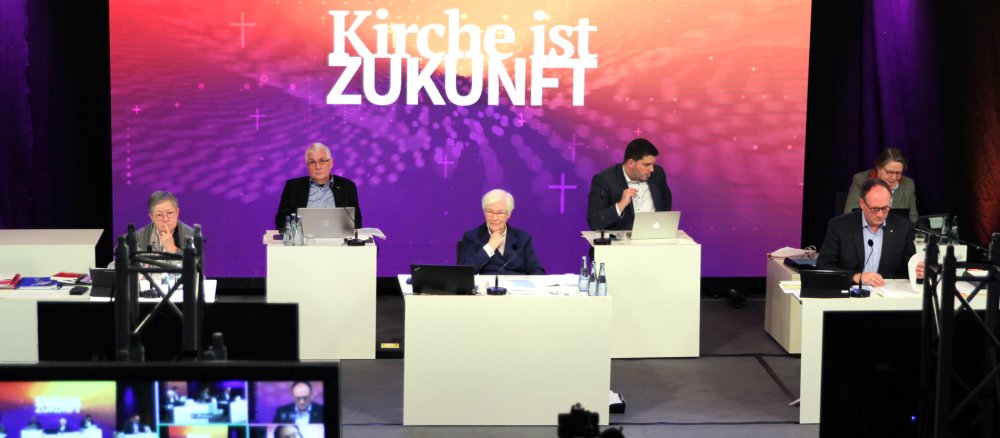 Drei Frauen und drei Männer sitzen vor einem Bildschirm mit der Aufschrift "Kirche ist Zukunft"
