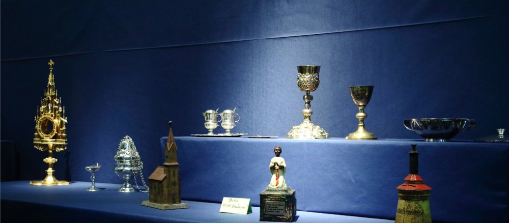 Kelche, Monstranzen und weitere Ausstellungsstücke vor blauem Hintergrund