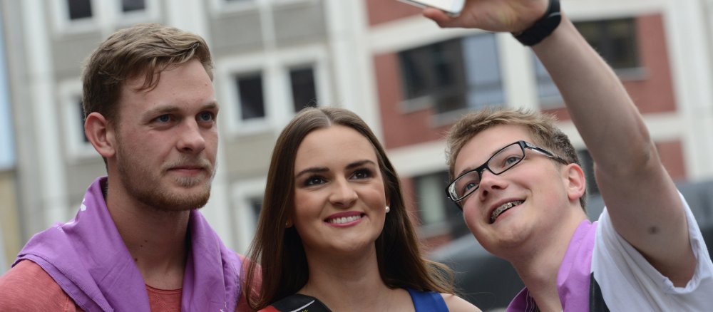 In der Mitte steht die dunkelhaarige Miss Germany, links und rechts zwei Junge Männer; einer davon macht ein mit seinem Smartphone über dem Kopf ein Selfie.