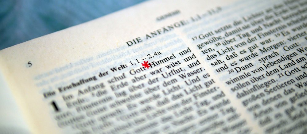 Eine bei Genesis 1,1 aufgeschlagene Bibel, beim Wort Gott ist ein rotes Sternchen ergänzt