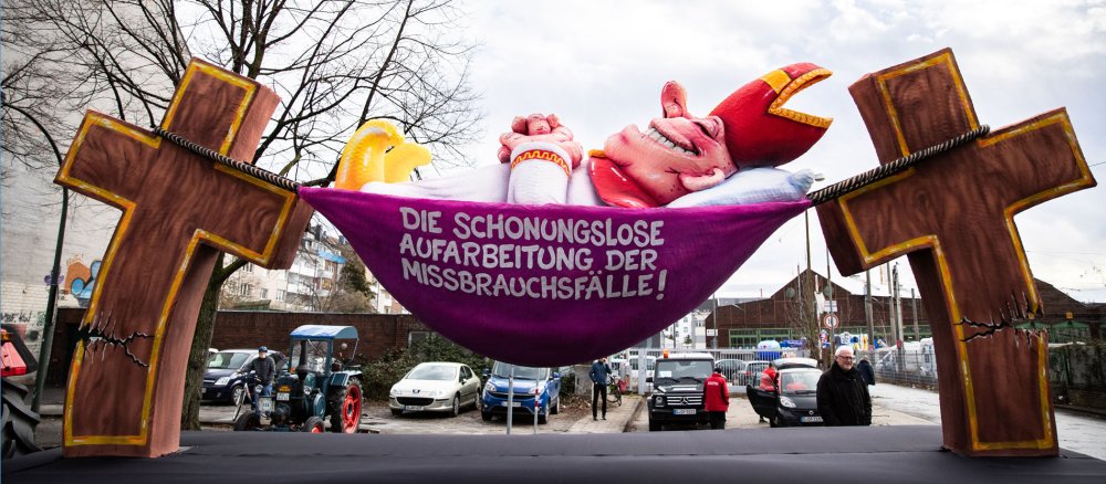 Ein politischer Mottowagen zur Aufarbeitung der kirchlichen Missbrauchsfälle mit der Figur eines kirchlichen Würdenträgers in einer Hängematte rollt vor dem Rosenmontagszug aus einer Halle