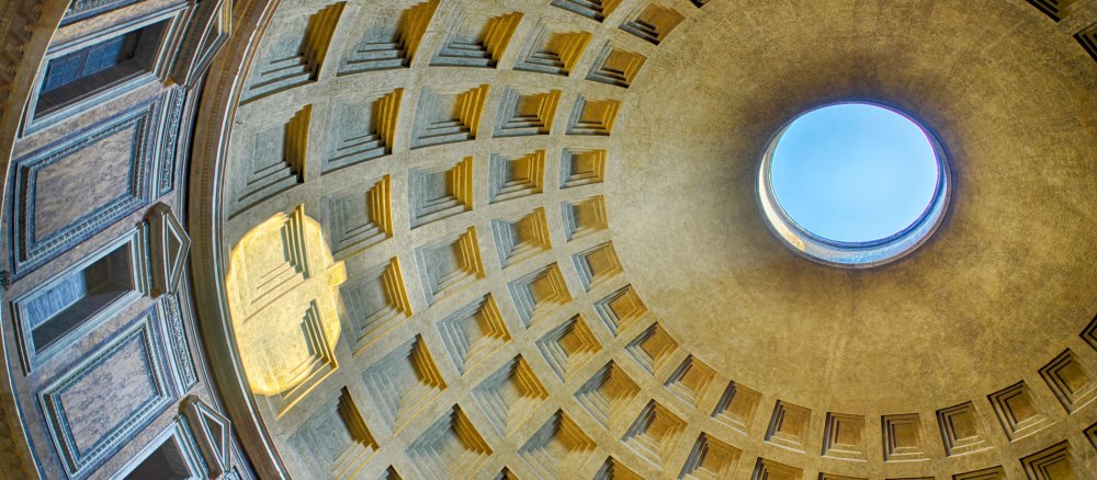 Man sieht von Innen auf die kasettenverdeckte Kuppel des Pantheons in Rom, das in der Mitte eine große runde Öffnung hat, durch die man den blauen Himmel sieht.