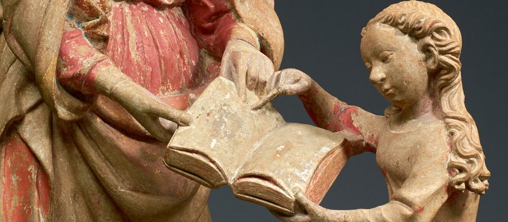 Maria zeigt mit einem Griffel auf die Stelle im Buch, die ihre Mutter Anna ihr weist.