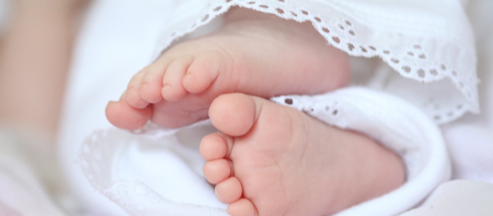 Ein Paar kleiner Babyfüße lugt unter weißem Kleid hervor.