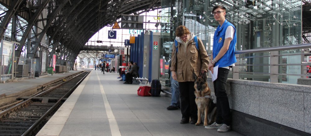 Der Kölner Hauptbahnhof. Am rechten Bildrand stehen eine Frau in einer braunen Jacke und ein junger Mann in einer Weste der Bahnhofsmission. Zwischen den beiden sitzt ein Schäferhund.
