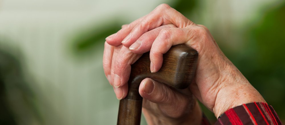Eine bewegungseingeschränkte Seniorin ruht sich aus und legt dabei die Hände auf ihren Gehstock.
