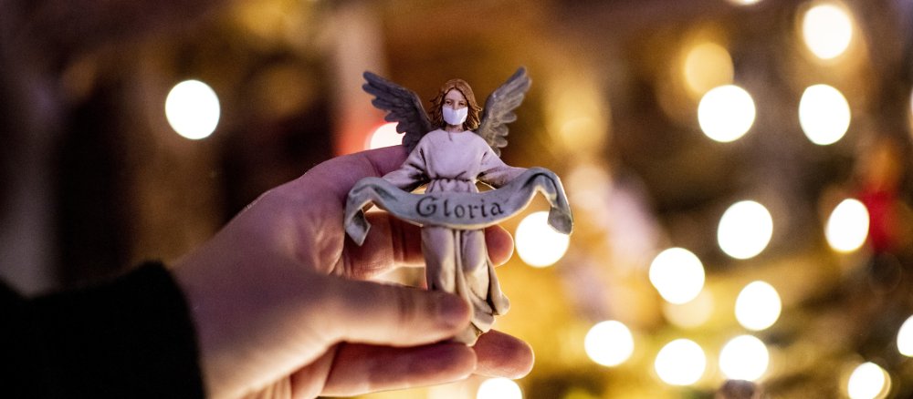 Eine Kinderhand hält die Krippenfigur des Engels Gabriel mit Mundschutz vor einer beleuchteten Krippe.