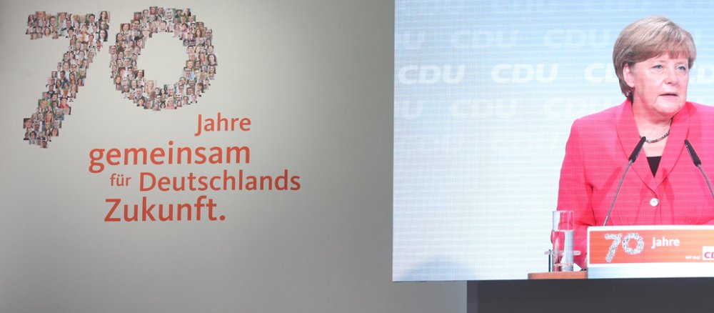 Angela Merkel ist auf einer Großbildleinwand zu sehen.
