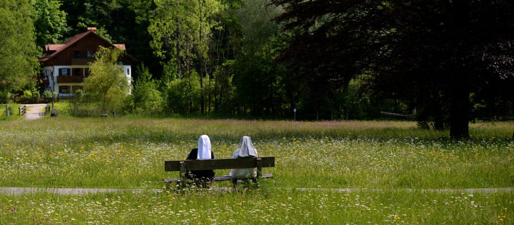 Zwei Ordensschwestern sitzen in Flüssen auf einer Bank vor einer Wiese.