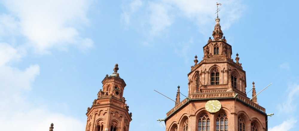 Blick von unten auf den Turm des rötlichen Mainzer Doms, blauer Himmel im Hintergrund