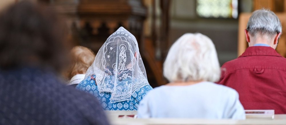 Rückansicht auf fünf Gottesdienstbesucher, eine Frau in der Mitte trägt ein weißes Kopftuch.