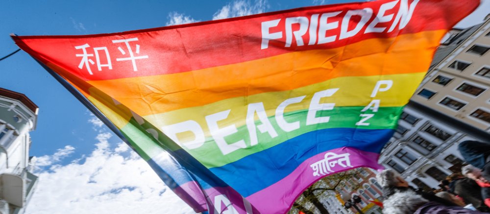Eine Regenbogenfahne, auf der das Wort Frieden in verschiedenen Sprachen geschrieben ist, weht im Wind.