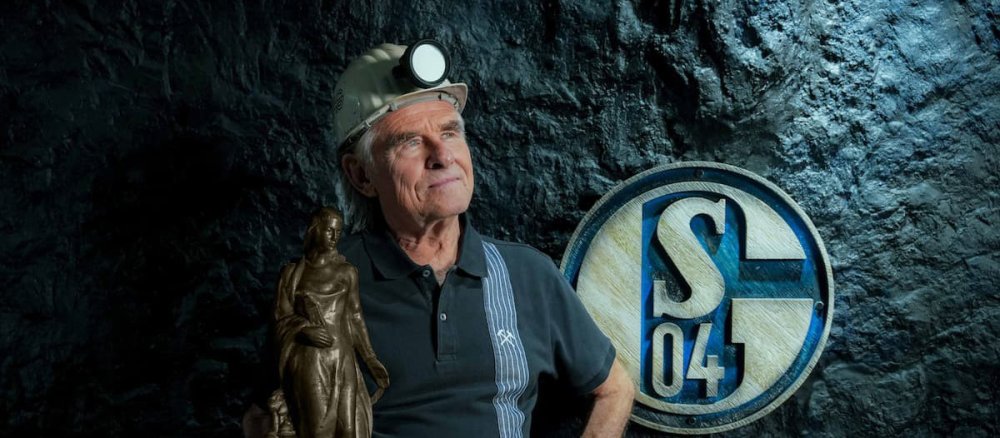 Ein Bergmann vor dem Schalke-Logo hält eine Statue der heiligen Barbara in den Händen