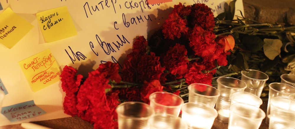 Blumen und Kerzen liegen auf dem Boden vor plakaten mit kyrillischen Schriftzeichen.