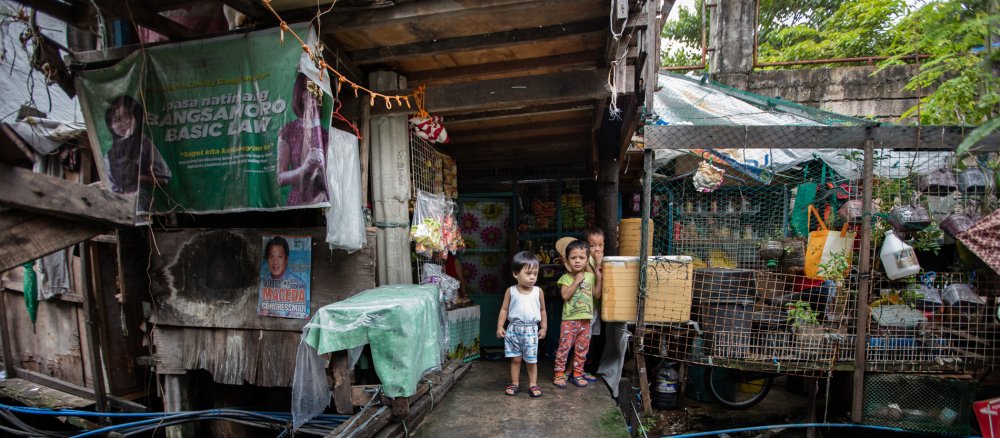 Drei Kinder stehen schüchtern vor einem Holzverschlag in einem philippinischen Slum
