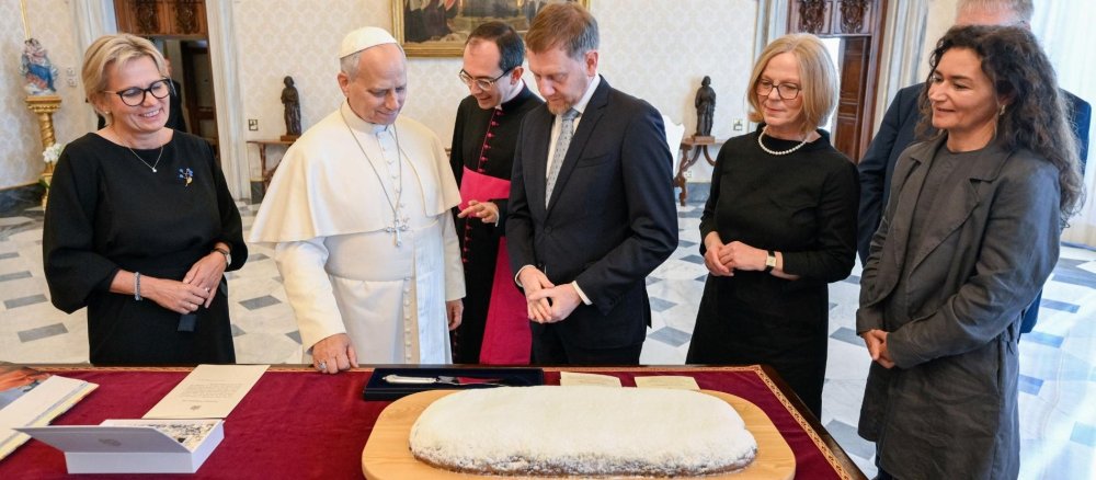 Papst Leo XIV. und Ministerpräsident Michael Kretschmer stehen zusammen mit anderen Menschen vor einem Tisch, auf dem ein großer Christstollen liegt