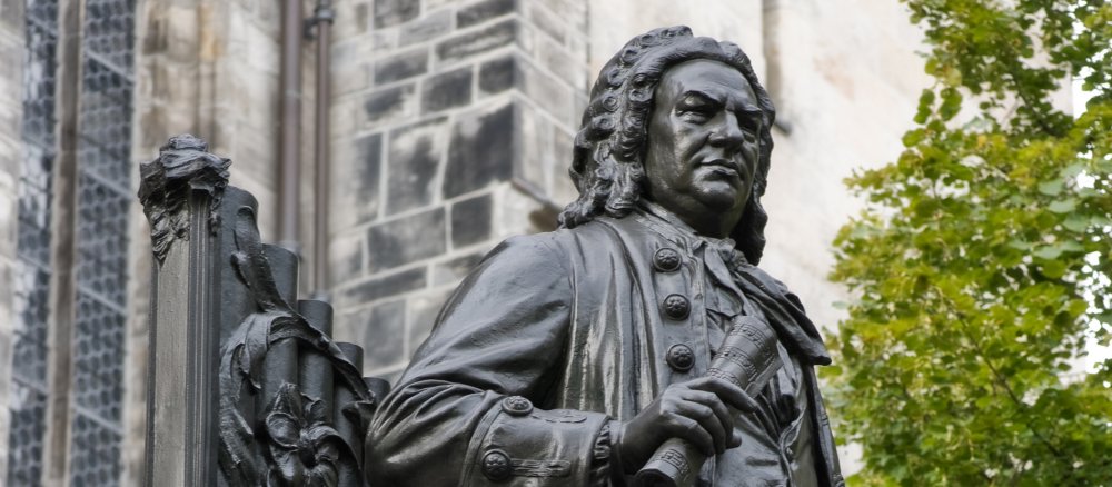 Bach trägt eine für damals übliche Perücke, lehnt sich an Orgelpfeifen an und hält in seiner rechten Hand zusammengerollte Noten.