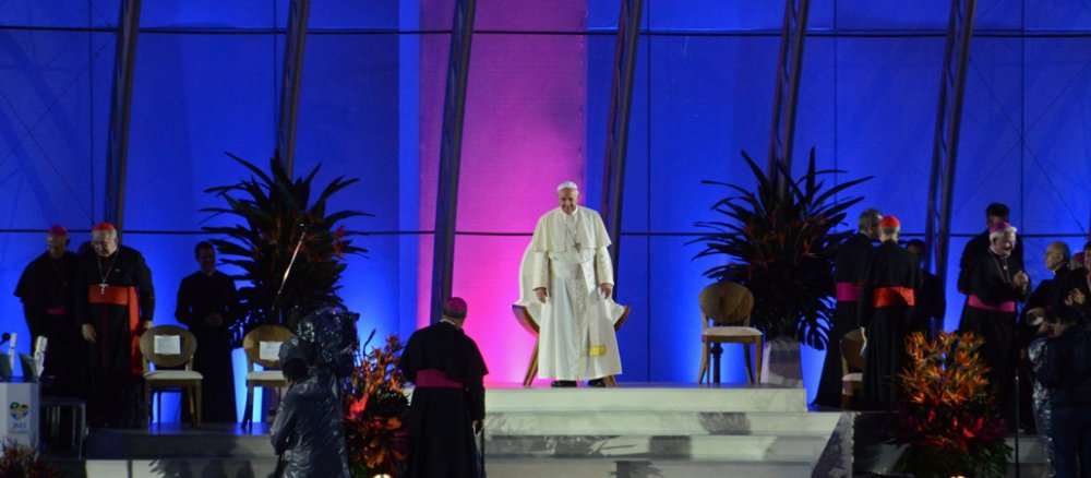 Der Papst steht aud einer Bühne, die hinter ihm blau und lila erleuchtet ist. Neben ihm stehen Bischöfe und Kardinäle.