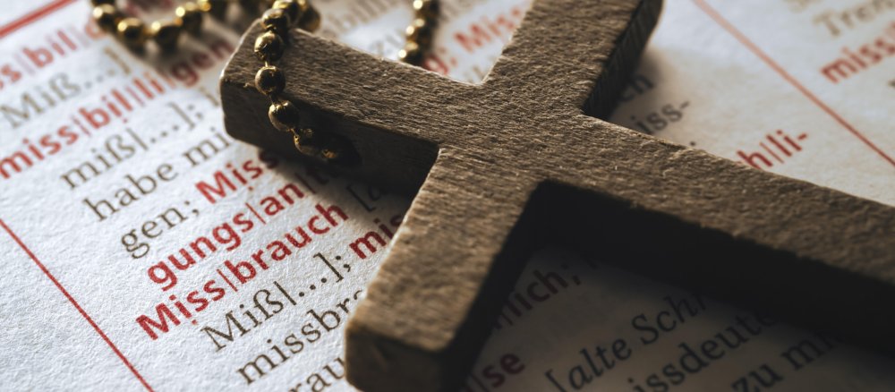 Ein Kreuz liegt auf einem Wörterbuch mit dem Stichwort "Missbrauch"
