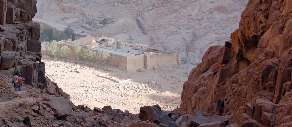 Durch eine riesige rötliche Felsspalte sieht man auf das hochummauerte Katharinenkloster in der Sinai-Wüste.