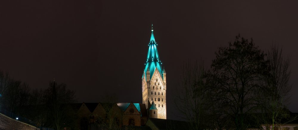 The tower of Paderborn Cathedral is illuminated in colour against a dark night sky.