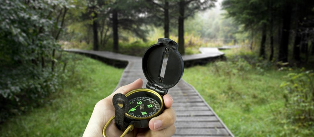 Eine Hand hält einen Kompass, weiter vorne gabelt sich der Weg aus Holzplanken.