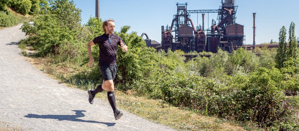 Pater Tobias Breer läuft auf einem Weg durch den Landschaftpark Nord in Duisburg, im Hintergrund sieht man ehemalige Industrieanlagen.