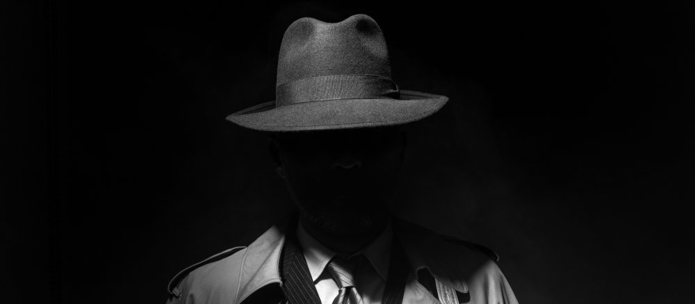 Ein Mann mit Hut und Trenchcoat verbirgt sein Gesicht im Schatten.