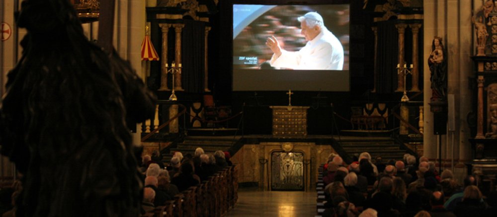 Hunderte Menschen sitzen in der abgedunkelten Kirche und schauen auf die viermal drei Meter große Leinwand.