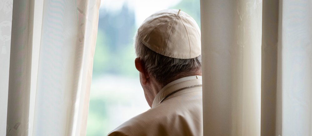 Papst Franziskus steht hinter einem Vorhang und guckt aus einem Fenster