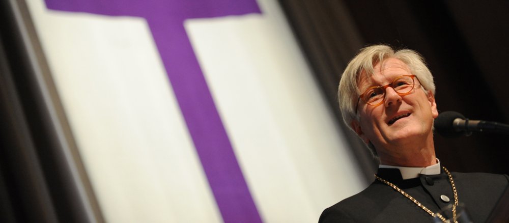 Der evangelische Landesbischof in Bayern, Heinrich Bedford-Strohm hält eine Rede, im Hintergrund ein violettes Kreuz.