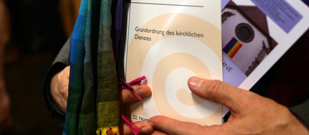 Ein Mann mit einem Regenbogenschal hält eine Ausgabe der Grundordnung in den Händen.